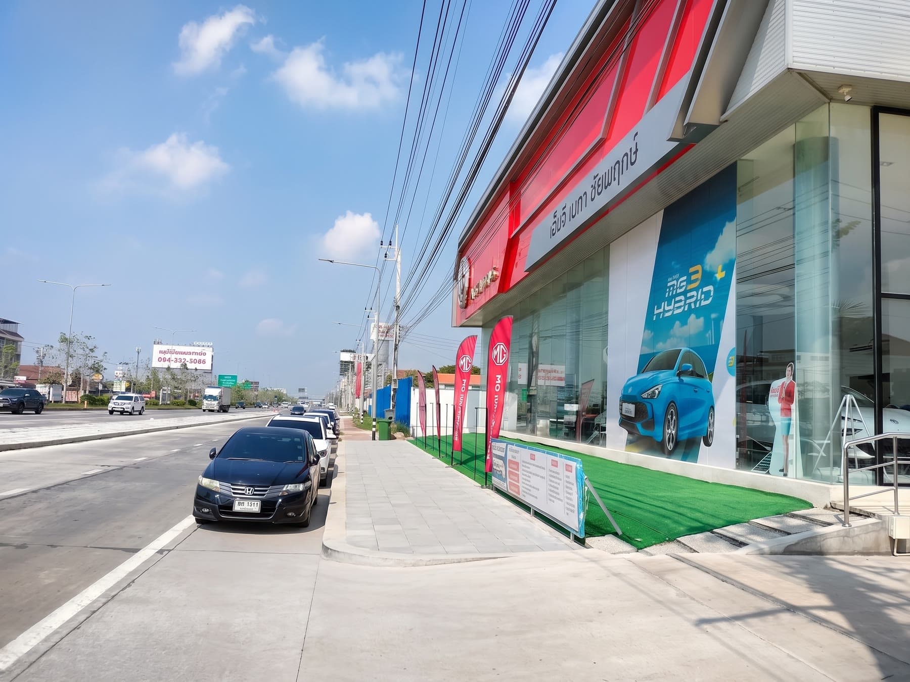 หน้าโชว์รูมมุมยาวริมถนน มีธง MG เรียงหน้าร้าน และป้าย MG3 Hybrid+ ติดที่กระจก